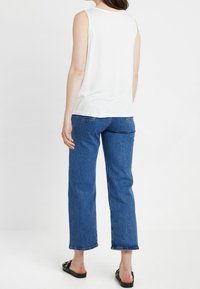 Biały top bez rękawów z teksturowanymi detalami na ramionach, zestawiony z niebieskimi jeansami o średnim stanie i prostych nogawkach oraz czarnymi klapkami.