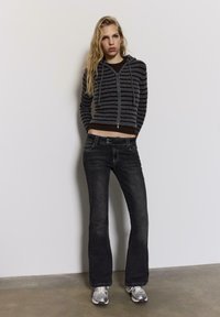 Sivo temno rjav kapucar s črtami, z zadrgo in oprijetim pasom, kombiniran z črnimi razširjenimi kavbojkami in srebrnimi supergami na betonski podlagi.