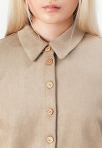 Beige collared shirt made of soft fabric, featuring five wooden buttons down the front and a smooth texture. Simple and elegant design.