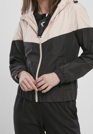 Femme portant une veste beige et noire zippée, tenant ses mains ensemble, avec des lunettes de soleil noires accrochées à la fermeture éclair de la veste.