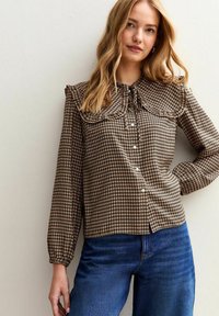 Brun rutigt blus med en stor volangkrage, långa ärmar och knäppning framtill; har texturerad tyg och en avslappnad passform.