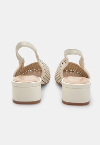 Sandalias de tacón beige con un diseño perforado, correa ajustable en el tobillo y una plantilla suave y acolchada. El tacón tiene forma de bloque sólido.