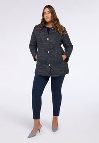 Marineblauer strukturierter Mantel mit pinken und weißen Sprenkeln, der einen hohen Kragen, große Knöpfe und Seitentaschen hat. Trägt kombiniert mit marineblauen Leggings und grauen Absätzen.