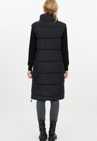 Lång svart pufferväst med hög krage, horisontell quiltning och dragsko nederkant, bärs över en svart långärmad topp och grå byxor.
