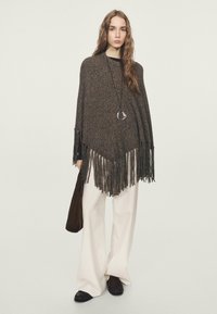 Poncho marrone lavorato a maglia con orlo con frange, indossato sopra una maglia nera, abbinato a pantaloni bianchi a gamba larga e scarpe nere. Accessoriato con una collana con ciondolo.