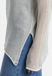 Weißes, geripptes Strickoberteil mit langen Ärmeln und seitlichen Schlitzern, strukturiert mit dünnen vertikalen Streifen. Kombiniert mit denimblauen Hosen.