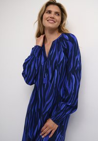 Sininen ja tummansininen mekko, jossa on rohkea, aaltoileva kuvio, pitkät hihat joustavilla hihansuilla ja halkiolla varustettu kaulus, jossa on kolme nappia.