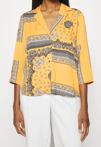 Camicia a maniche corte, con colletto, in tessuto arancione brillante decorata con motivi paisley neri e bianchi e strisce; vestibilità comoda e texture leggera.