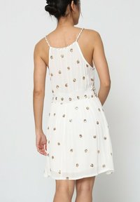 Robe sans manches blanche avec une taille cintrée, ornée de pois dorés et d'une jupe fluide. Tissu doux et texturé avec un léger scintillement.