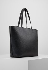 Svart tote-bag laget av teksturert syntetisk materiale med et preget logo-mønster. Den har høye, glatte dobbeltremmer og en rektangulær form.
