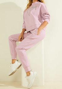 Světlé růžové loungewear oblečení zahrnující zkrácenou mikinu s potiskem loga a odpovídající jogger kalhoty s bočními kapsami, doplněné bílými teniskami.