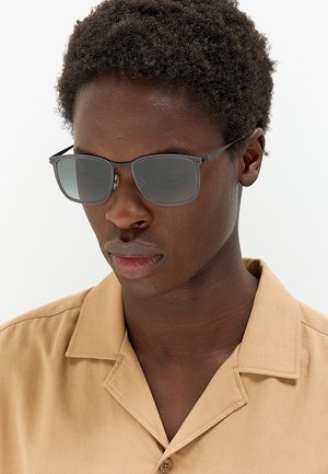 Giovane con capelli corti e ricci, che indossa occhiali da sole rettangolari scuri e una camicia beige con bottoni su uno sfondo semplice.