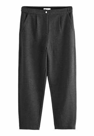 Femme portant un pull violet, un pantalon large foncé, des talons noirs pointus, tenant un grand sac à main noir à l'intérieur près de grandes fenêtres.