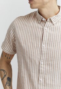Chandail à manches courtes, à boutons, de couleur brun clair avec des rayures verticales blanches. Fabriqué en tissu texturé avec une finition douce et un col rabattable.
