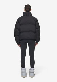 Svart puffjacka med hög krage, texturerat tyg, logotyp på ryggen, i kombination med svarta leggings och grå snöskor.