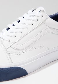 Sneaker bianco in pelle con suola in gomma blu testurizzata, lacci bianchi e cucitura laterale. Presenta un design minimalista e un profilo piatto.