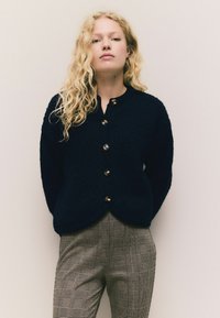 Ung kvinde med langt, krøllet, blond hår står foran en ensfarvet baggrund, iført en sort, knappet cardigan og ternede bukser.