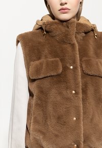 Brauner Kunstfell-Weste mit einer gepolsterten beige Kapuze. Verfügt über zwei Fronttaschen und goldfarbene Beschläge an den Zugbändern. Weicher Stoff.