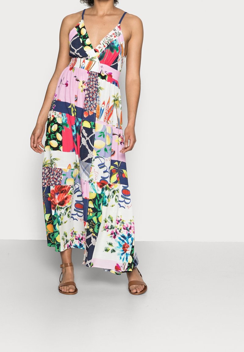 Femme portant une robe longue sans manches en patchwork multicolore avec des motifs floraux et abstraits, associée à des sandales à lanière à la cheville couleur tan.