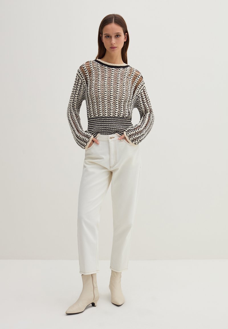Schwarz-weiß gemusterter Strickpullover mit langen Ärmeln, kombiniert mit weißen, hochgeschnittenen Hosen und beigen spitzen Knöchelschuhen.