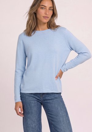 Femme aux cheveux châtain clair, portant un pull bleu clair à manches longues et un jean bleu, debout sur un fond clair uni.