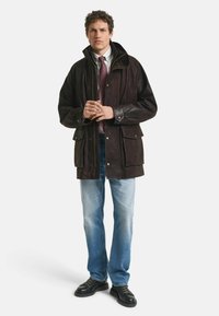 Bruine leren jas met een hoge kraag, meerdere zakken en een rits. Gecombineerd met lichtblauwe jeans en zwarte schoenen.
