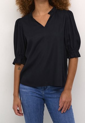 Femme portant un chemisier noir à manches courtes volantées et encolure à volants, associé à un jean bleu, debout devant un fond uni.