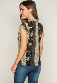 Blouse fleurie à manches courtes, avec un fond noir ornée de motifs jaunes et bleus, un col texturé et des sections de tissu contrastantes.