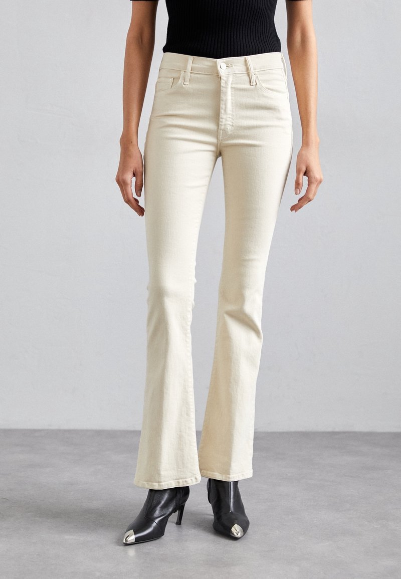 Jean évasés couleur crème en denim, dotés d'une taille haute et d'un design à cinq poches, associés à des bottes à talons noires.