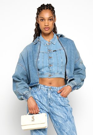 Donna con capelli intrecciati che indossa una giacca di jeans a strati e una camicia di jeans corta, jeans acidati, che tiene una piccola borsa bianca.