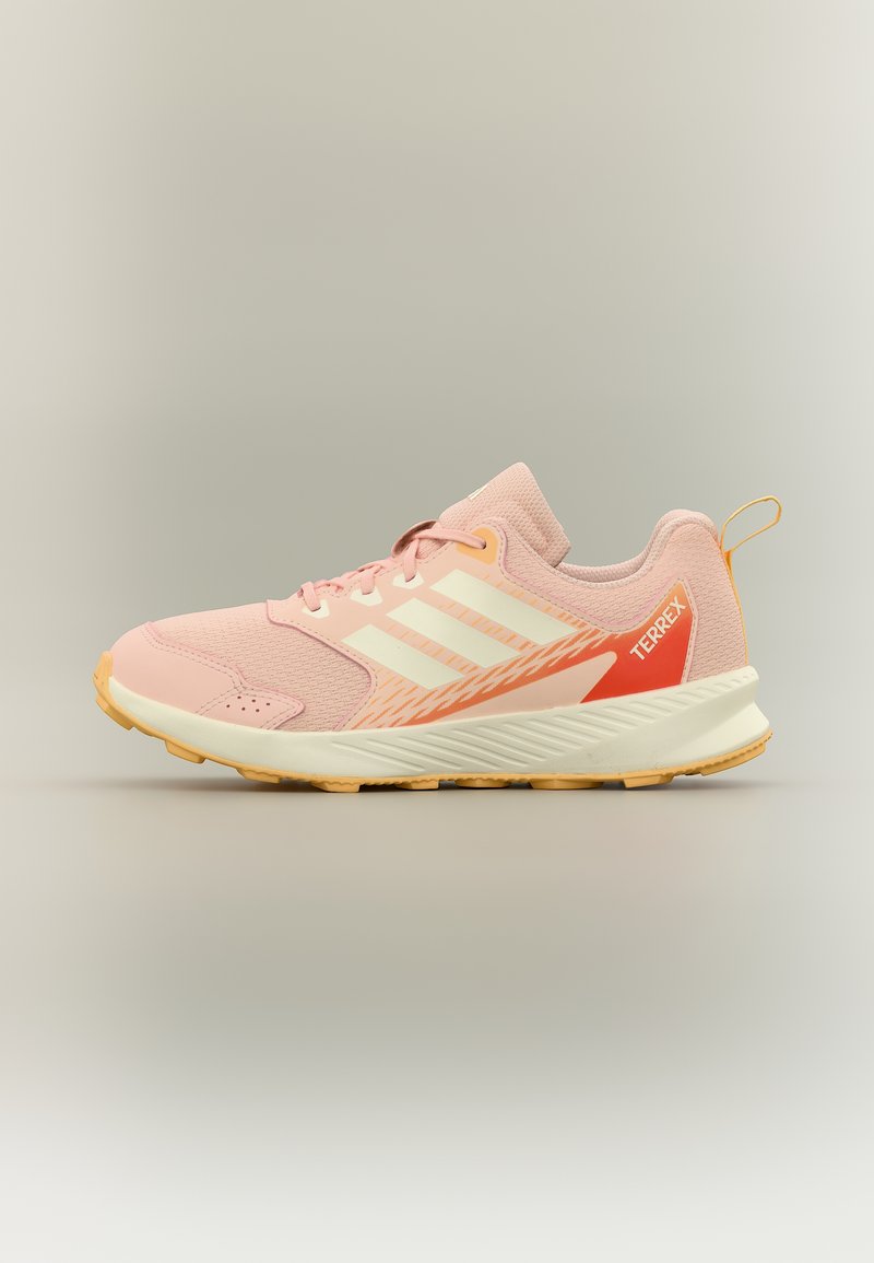 Απαλό ροζ αθλητικό παπούτσι Adidas Terrex με λευκές ρίγες, πορτοκαλί λεπτομέρεια στη φτέρνα και καφέ λάστιχο σόλας, εμφανίζεται σε προφίλ πλευράς σε ουδέτερο φόντο.