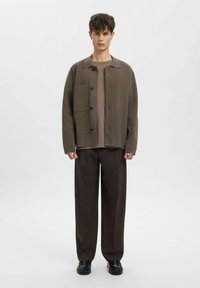 Nadměrně velká olivově zelená bunda se dvěma předními kapsami, černými knoflíky a béžovým tričkem s kulatým výstřihem pod ní, doplněná hnědými kalhotami s širokými nohavicemi.