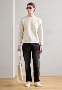 Felpa beige con una piccola tasca sul petto, jeans grigio scuro, sneakers bianche e una borsa tote color crema. Modello che indossa occhiali da sole.