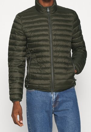 Mann trägt dunkelgrüne, gesteppte Jacke mit Reißverschluss und hohem Kragen sowie blaue Jeans, Hände entspannt an den Seiten.