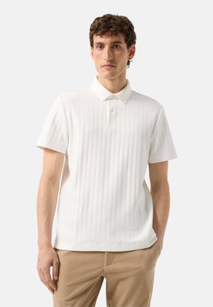 Mann trägt ein weißes kurzärmeliges, geripptes Poloshirt und beige Hosen, steht vor einem schlichten hellen Hintergrund.