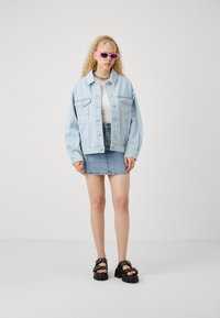 Chaqueta oversized de denim azul claro, top blanco de canalé, falda de denim azul ajustada y sandalias negras con tachuelas. Gafas de sol rosas como acento.