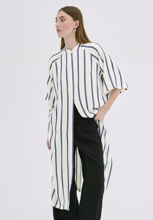 Lange gestreepte shirt in crème en marineblauw, met een knoopsluiting aan de voorkant, brede mouwen, een relaxte pasvorm en zijspleten. Gecombineerd met zwarte broek.