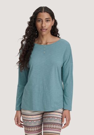 Frau mit langen lockigen Haaren, die ein blaues langärmliges Oberteil und gemusterte Leggings trägt, steht vor einem schlichten Hintergrund.