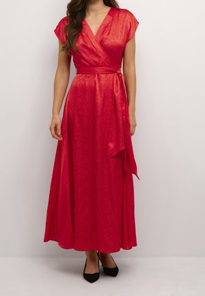 Vrouw die een rode wikkeljurk met korte mouwen en een ceintuur draagt, gecombineerd met zwarte puntige hakken, staand tegen een effen achtergrond.