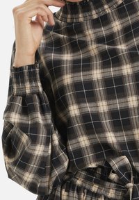 Black and beige plaid blouse with smocked high collar and puff sleeves. The fabric is smooth with a soft sheen, featuring a checked pattern.