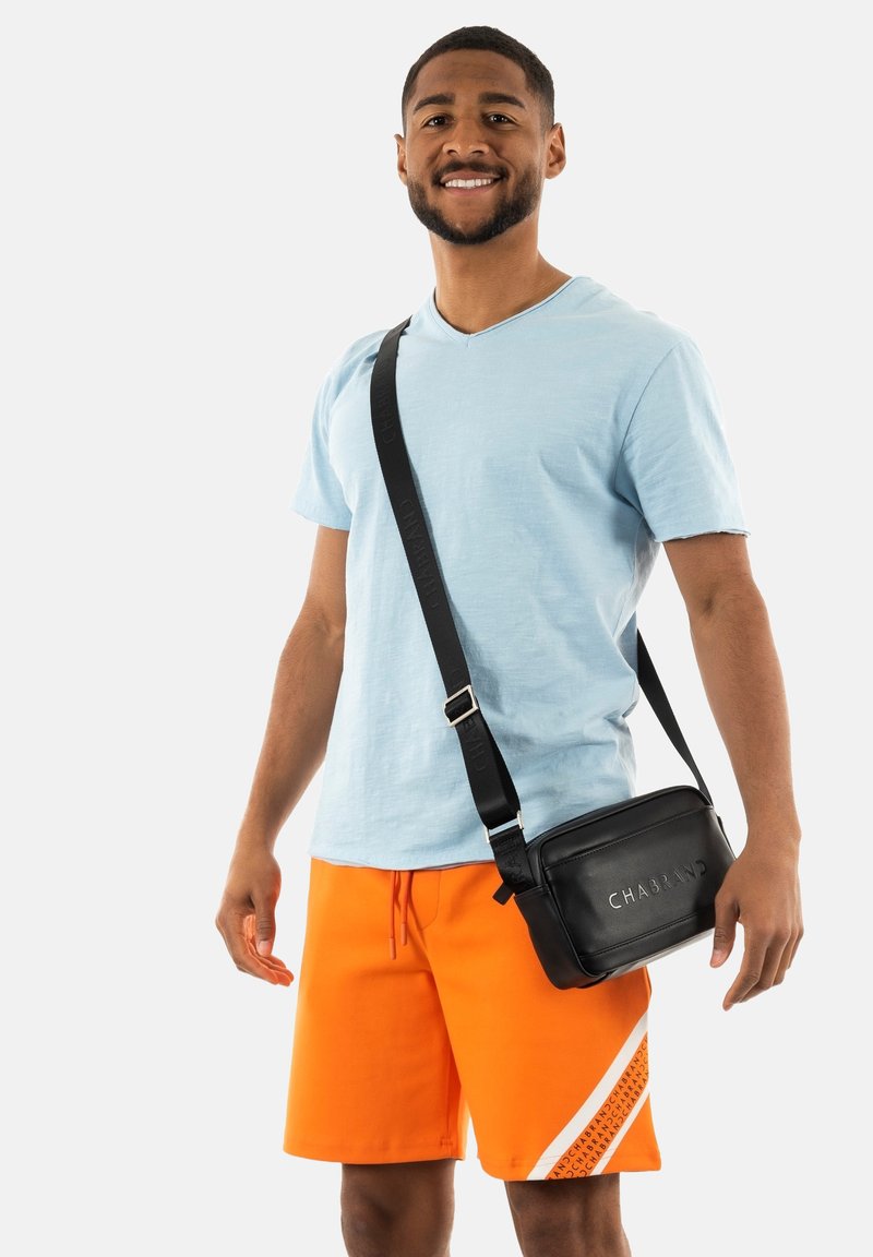 Borsa a tracolla nera con "CHABRAND" impressa, indossata sopra una maglietta a V di colore blu chiaro e pantaloni corti arancioni con un motivo bianco sull'orlo.