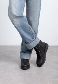 Botas negras hasta el tobillo con punta redondeada, cuero texturizado y cordones. Combinadas con unos pantalones vaqueros claros, holgados y remangados. Fondo de suelo gris.