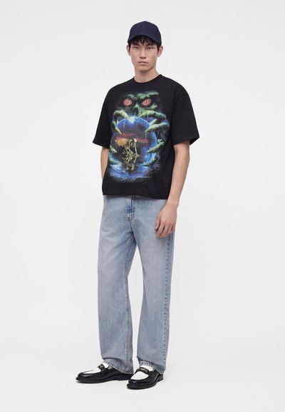 Νέος άνδρας που φοράει μαύρο γραφικό T-shirt με χέρια και μάτια τεράτων, ανοιχτό μπλε τζιν, μαύρα και λευκά παπούτσια και σκούφο μπέιζμπολ.