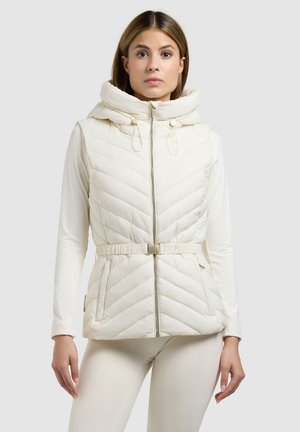 Žena v bílém bezrukávovém prošívaném vestu s kapucí, předním zipem a páskem, oblečená přes dlouhý bílý top a bílé kalhoty, stojící na šedém pozadí.