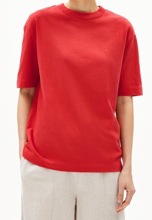 Personne portant un t-shirt rouge uni à manches courtes et un pantalon beige clair, les mains détendues le long du corps, devant un fond uni.