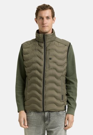 Man wearing olive green padded vest over matching long sleeve shirt and light gray jeans, standing against white background.