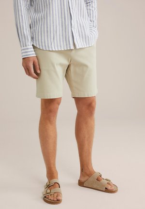 Helle beigen Shorts aus Baumwolle, kurz über dem Knie, kombiniert mit tanfarbenen Wildledersandalen mit verstellbaren Schnallen. Blau-weiß gestreiftes Hemd.