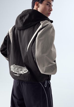 Mladić s kratkom tamnom kosom u crno-bež jakni s kapuljačom i grafičkim tekstom na leđima, gleda preko ramena.