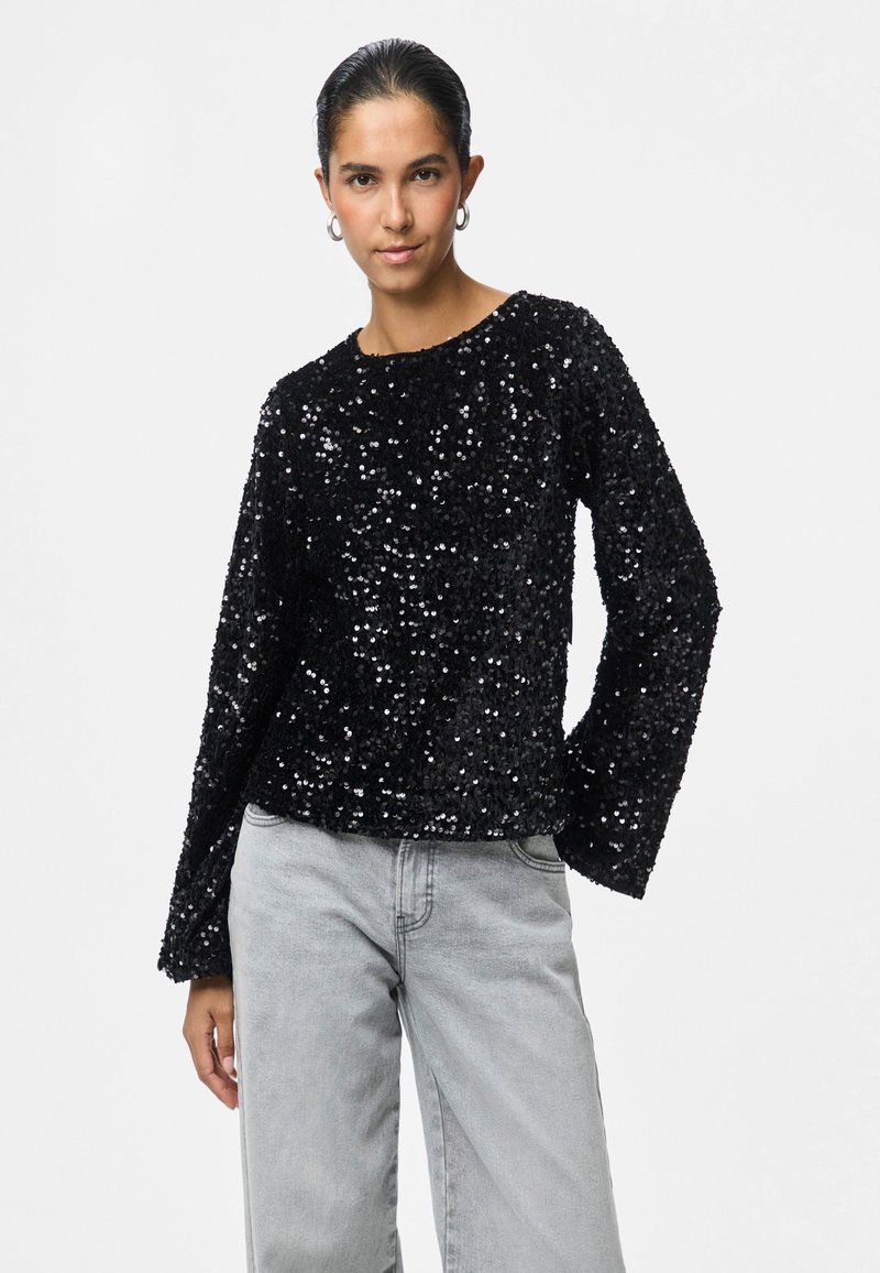 Mulher com cabelo escuro preso para trás, usa brincos de argola, uma blusa de manga longa preta com lantejoulas e jeans de cintura alta cinza claro, de pé com uma mão para trás.