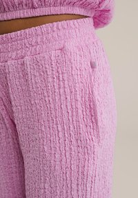 Roza teksturirane hlače iz gubanega blaga, ki imajo stransko žep in majhno oznako logotipa. Pas je elastičen za udobje.